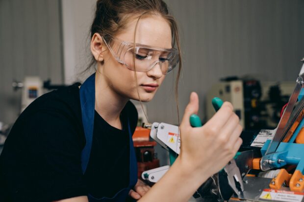Kobieta w stroju roboczym i okularach ochronnych pracuje ze śrubokrętem w warsztacie.