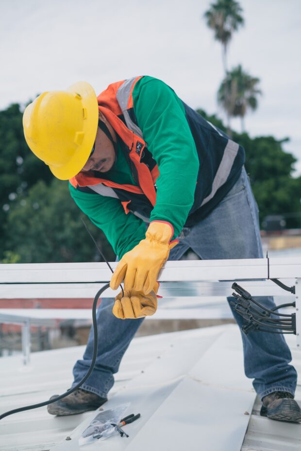 Serwisant w żółtym kasku i rękawicach ochronnych pracuje na blaszanym dachu z czarnym okablowaniem.